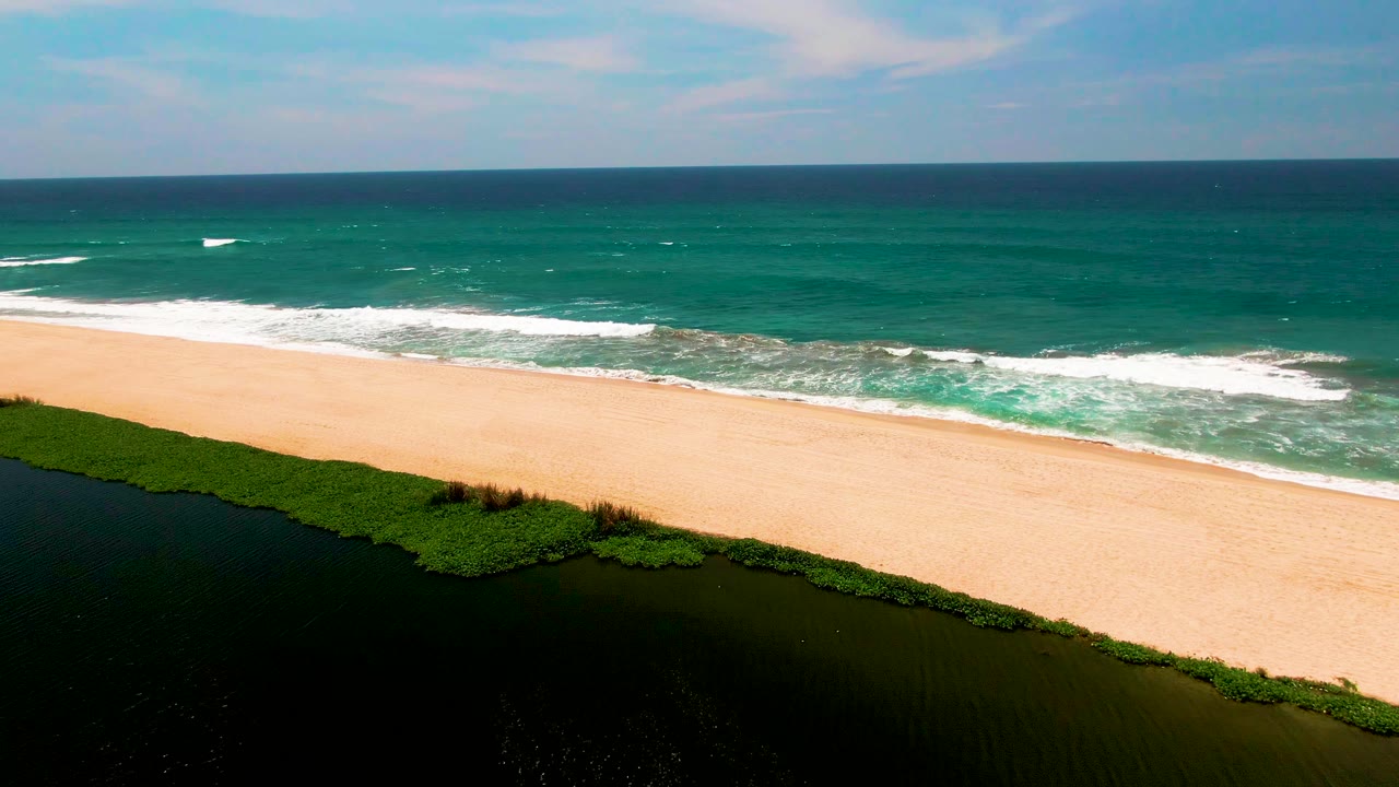 The San Jose del Cabo shoreline appears from above with lagoon vegetation at lower left, a clear band of sand, white surf moving toward shore, and no visible people.