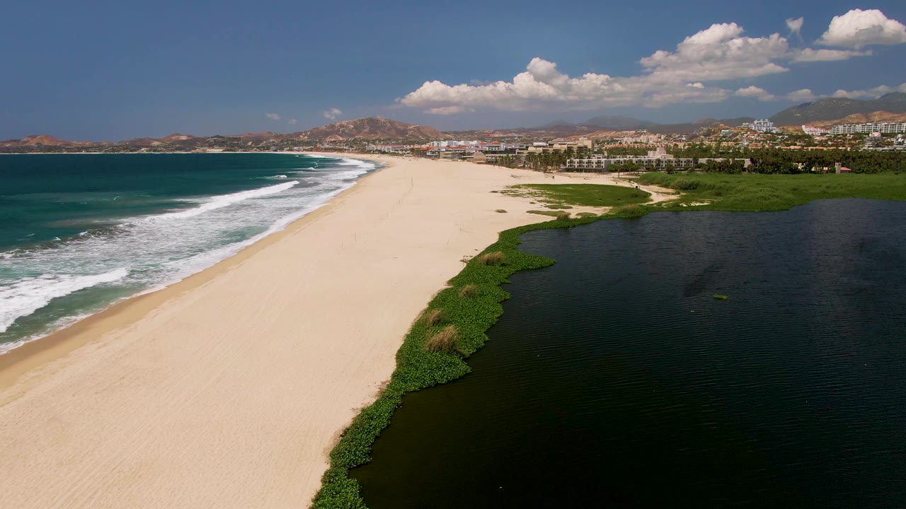 Aerial view in San Jose Del Cabo showing teal ocean surf on the left, pale sand through the center, a dark lagoon on the right, distant hills and buildings, and no visible people.