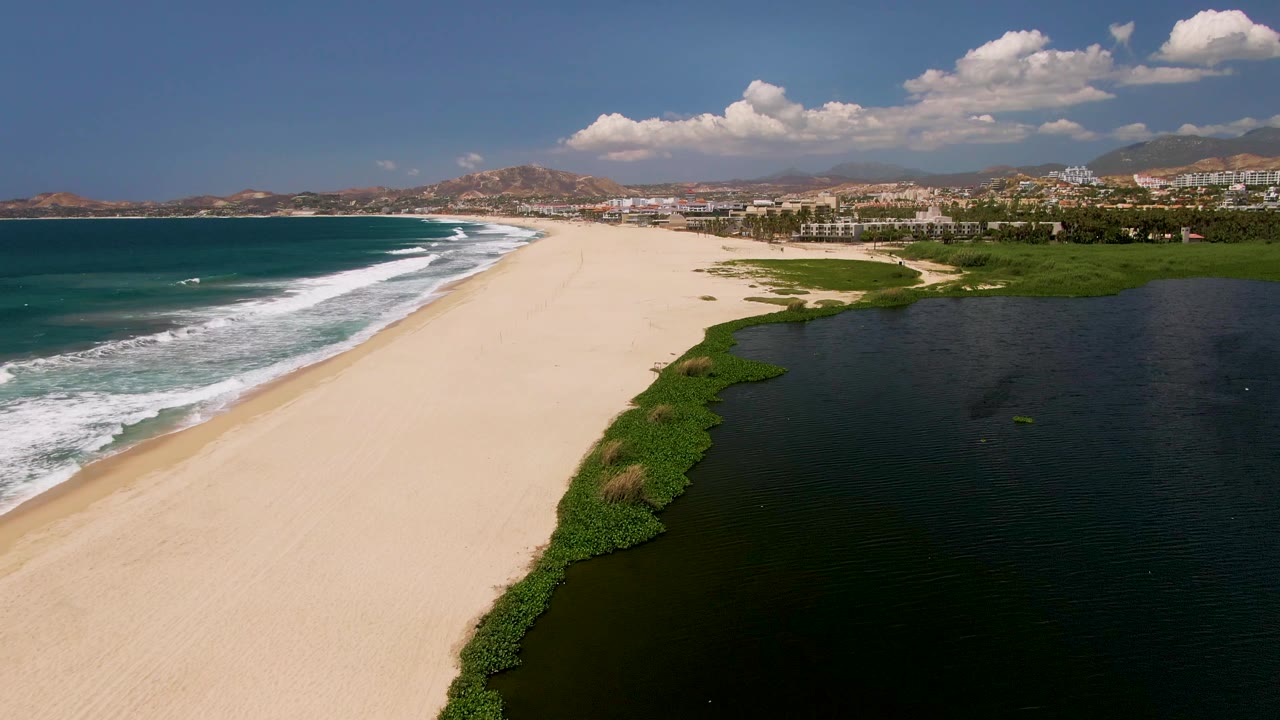 Aerial view in San Jose Del Cabo of a broad sand strip separating foamy ocean waves from the dark lagoon, with green reeds at the lagoon edge, distant buildings and hills, and no visible people.