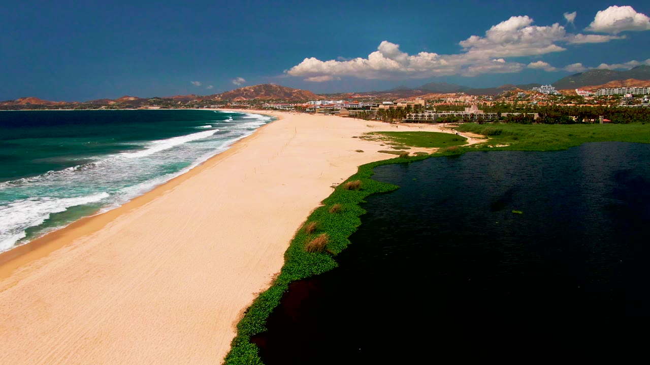 Wide beach aerial with surf lines, lagoon edge, coastal greenery, distant hills, and buildings.