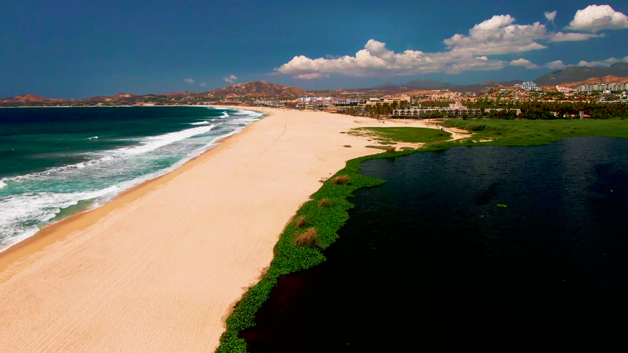 A high aerial view of San Jose del Cabo shows a broad empty beach between teal ocean waves and a dark lagoon, with green vegetation, low hills, scattered buildings, bright clouds, and no visible people.