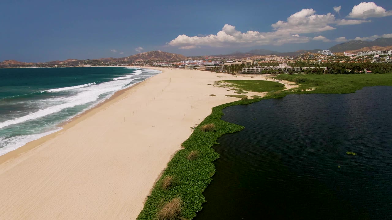 Aerial frame from San Jose del Cabo showing a wide sandy beach between blue ocean surf on the left and a dark lagoon edged with green vegetation on the right, with no visible people.