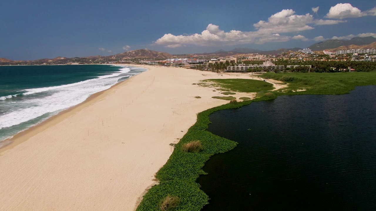 High aerial frame from San Jose del Cabo showing a pale beach between rolling ocean waves and a dark lagoon, with green vegetation, coastal buildings, dry hills, and no visible people.