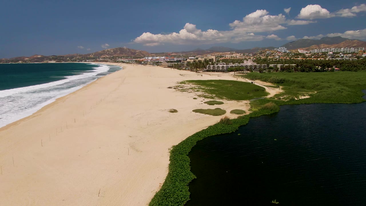 Aerial frame over San Jose Del Cabo beach showing ocean surf at left, pale sand through the center, a dark lagoon at lower right, distant buildings, hills, clouds, and no visible people.