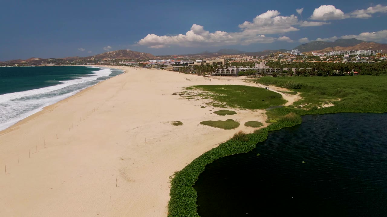 Aerial beach view with ocean waves, lagoon water, vegetation, and distant buildings.