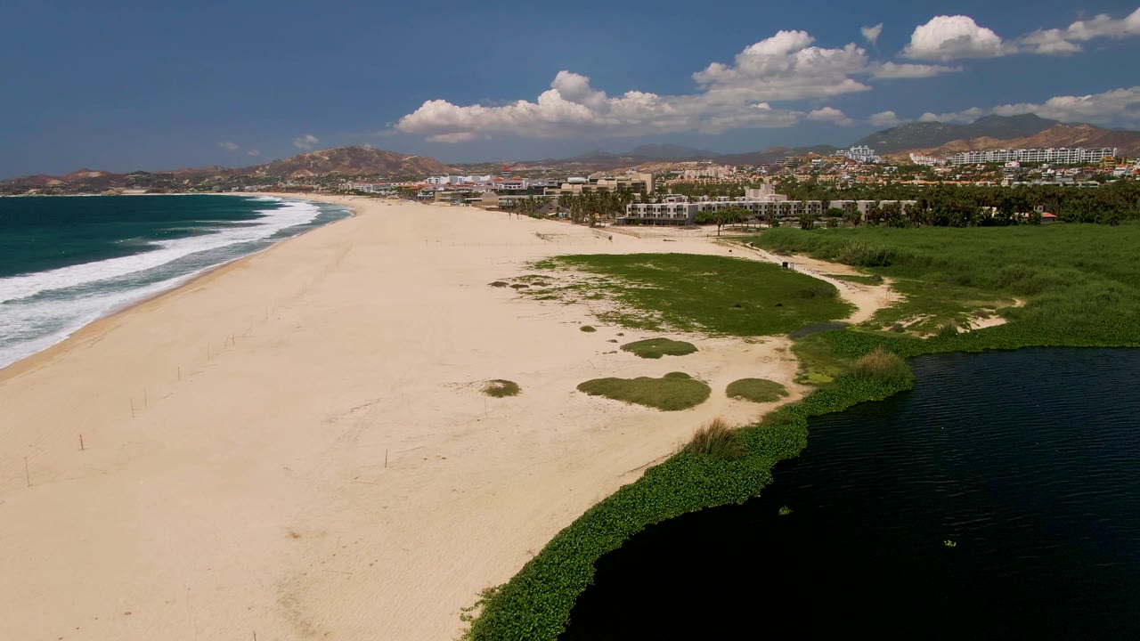At 0:02, the aerial view shows the surf line pushed along the left beach edge, a larger dark lagoon area in the lower right, scattered green growth on the sand, distant San Jose Del Cabo buildings, and no visible people.