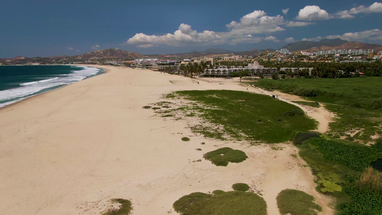 Aerial frame over a San Jose del Cabo beach with pale sand below, green grass patches to the right, surf rolling in from the left, inland buildings, distant mountains, blue sky, white clouds, and no visible people.