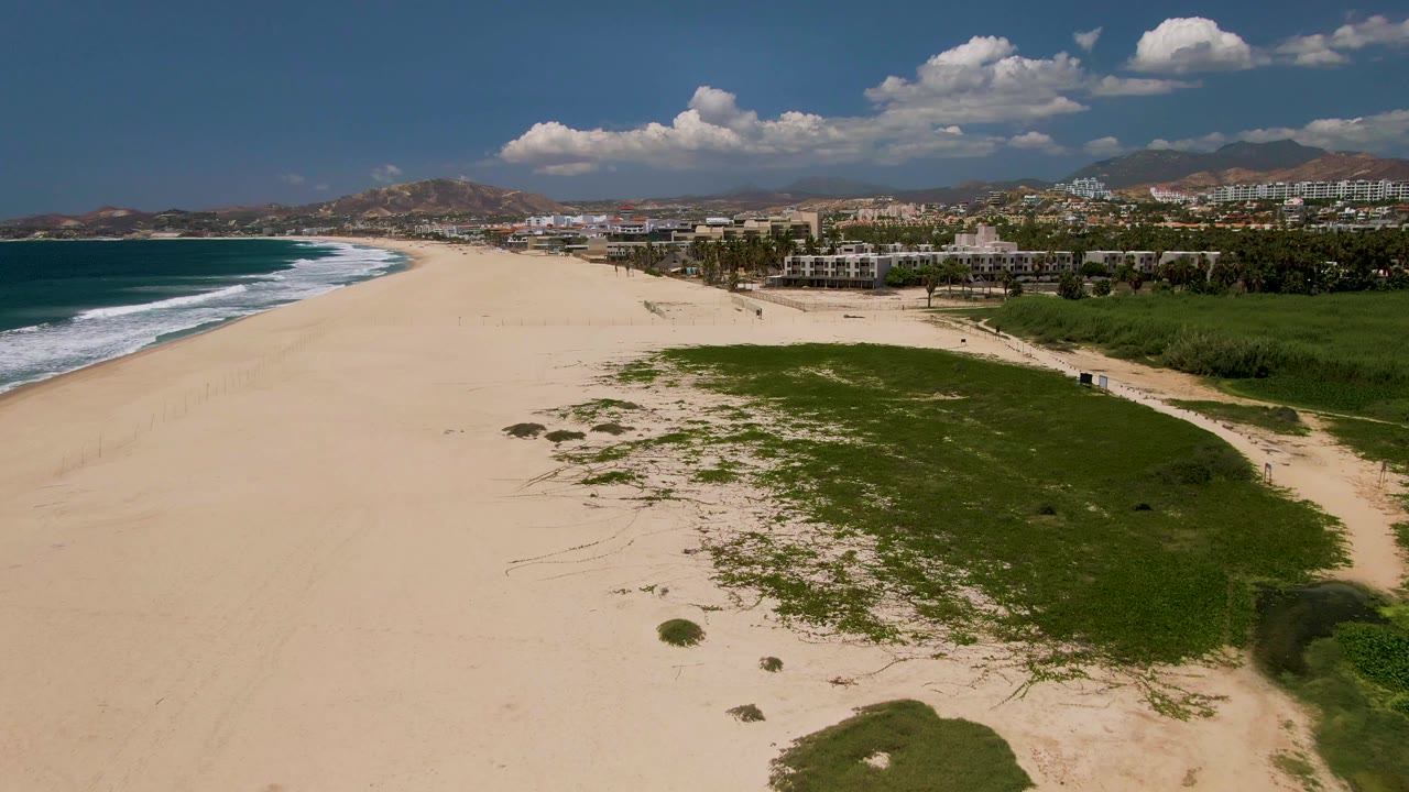 A high view of the San Jose del Cabo shoreline shows ocean water and white waves on the left, a large green grass patch over the sand on the right, low buildings inland, distant hills, and no visible people.