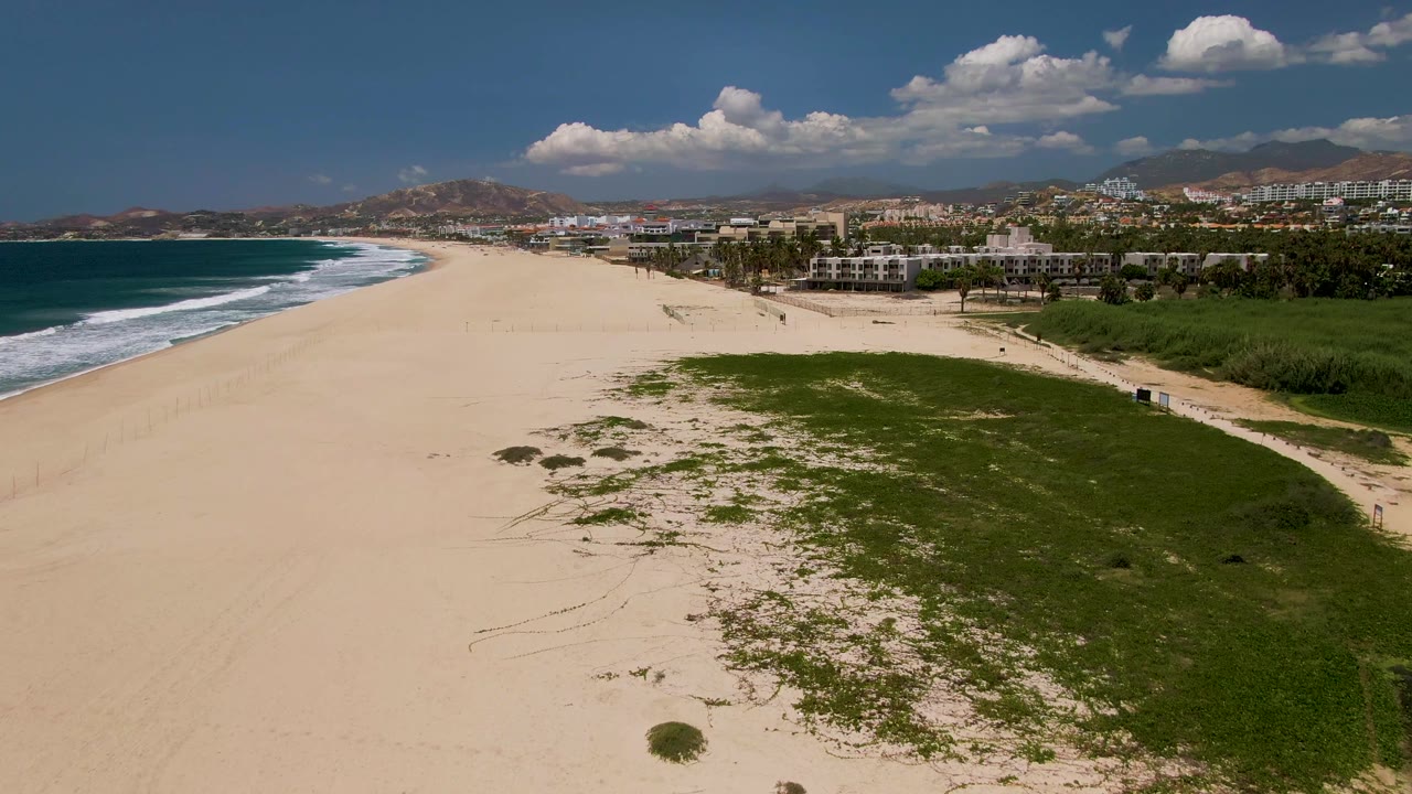 Aerial view along a sandy coastline with dark blue water, white surf, green beach grass, and inland mountains.