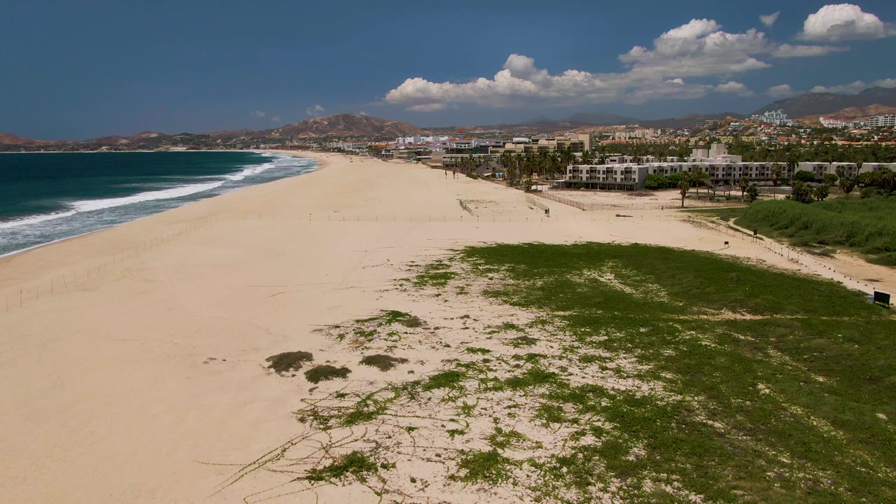 Wide aerial view of a San Jose del Cabo beach with ocean surf on the left, green dune vegetation in the foreground, beachfront buildings to the right, distant hills, and tiny people visible on the sand.