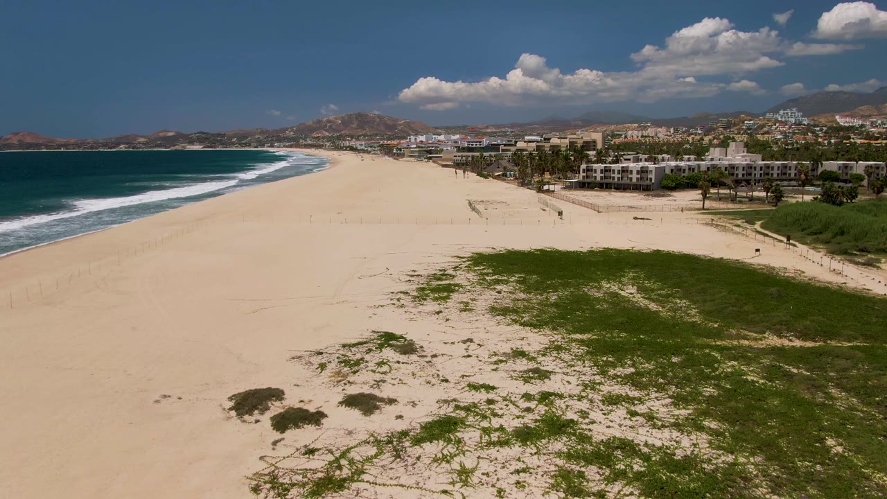 Aerial coastline view with broad sand, surf, dunes, and beachfront buildings.