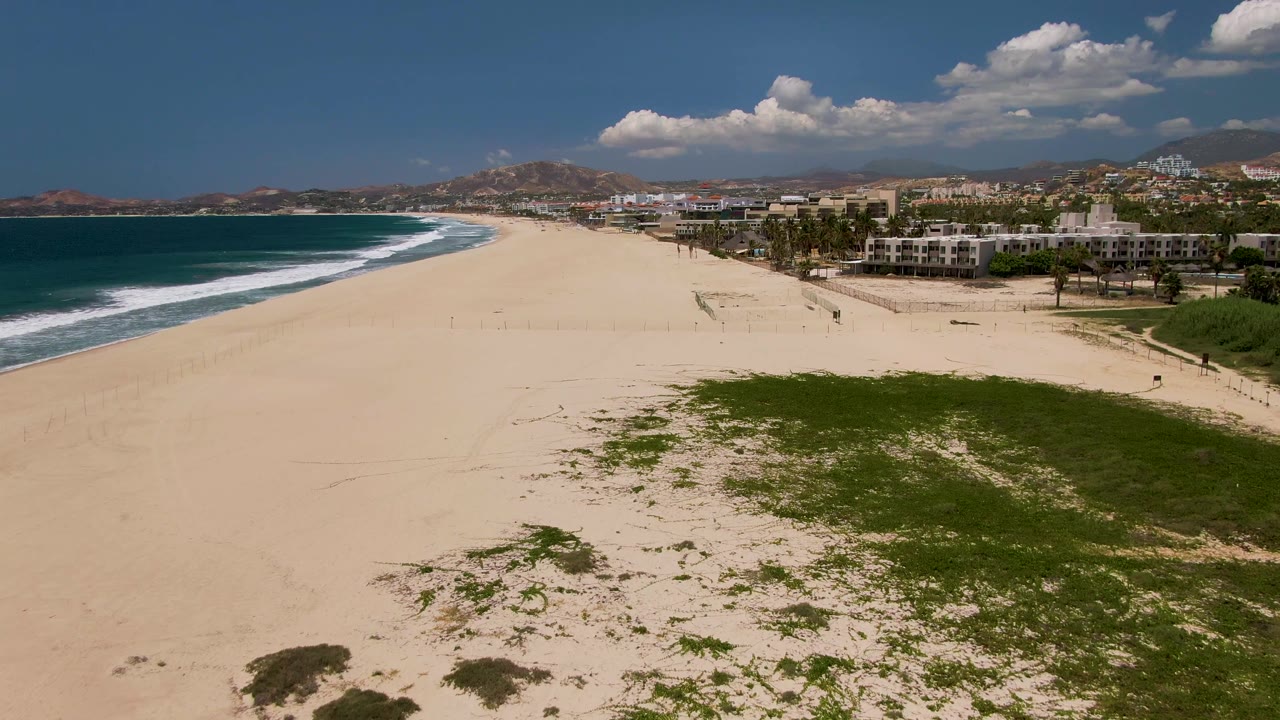 Sunny San Jose del Cabo aerial beach frame with turquoise ocean and white surf left, a large open sand area, green dune vegetation lower right, low buildings and palms along the beach edge, and small people visible.