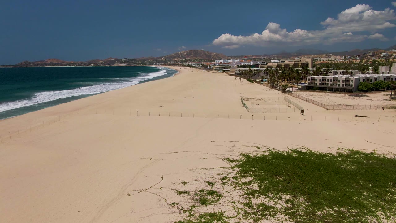 Aerial view of sandy beach, ocean waves, buildings, and distant hills.