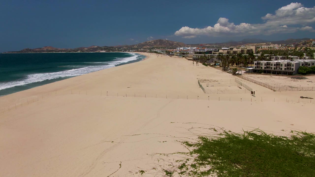 Aerial view over San Jose del Cabo shoreline with the surf curving across the left side, a large empty sand area, buildings beyond palms, and no visible people.