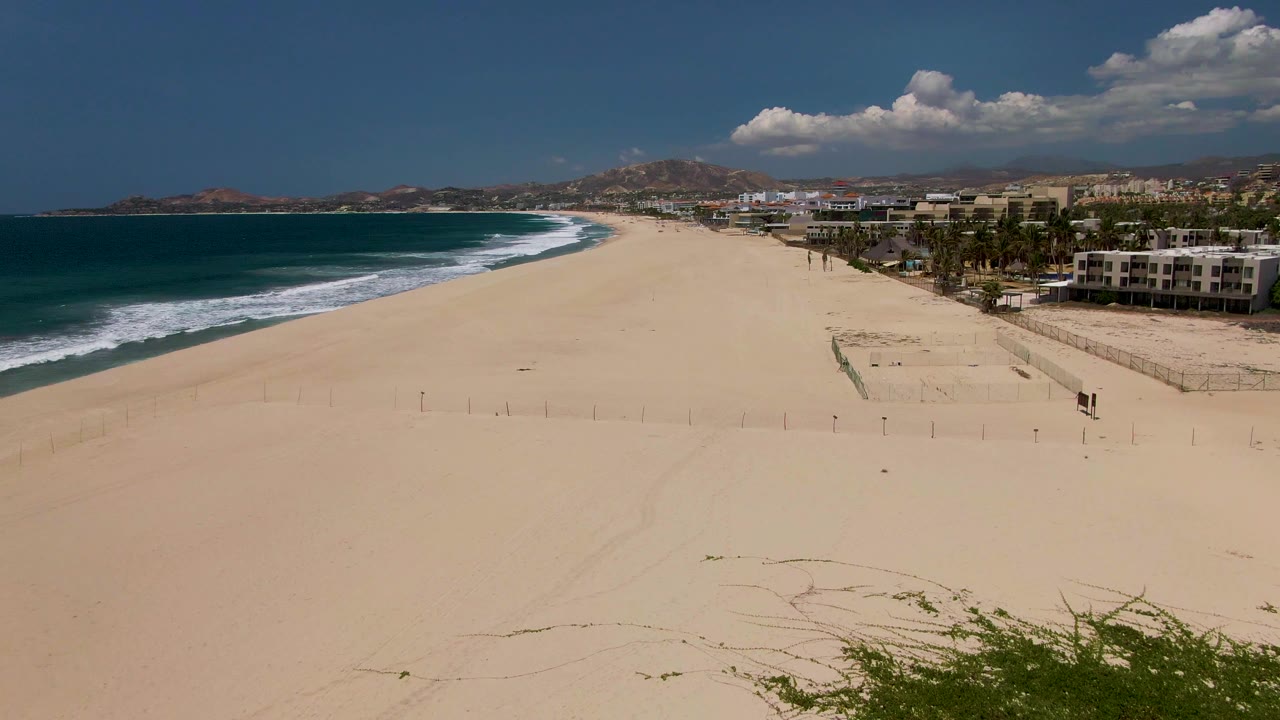 San Jose del Cabo beach seen from above with darker blue water to the left, bright sand stretching toward distant buildings, palm trees along the coast, and no visible people.