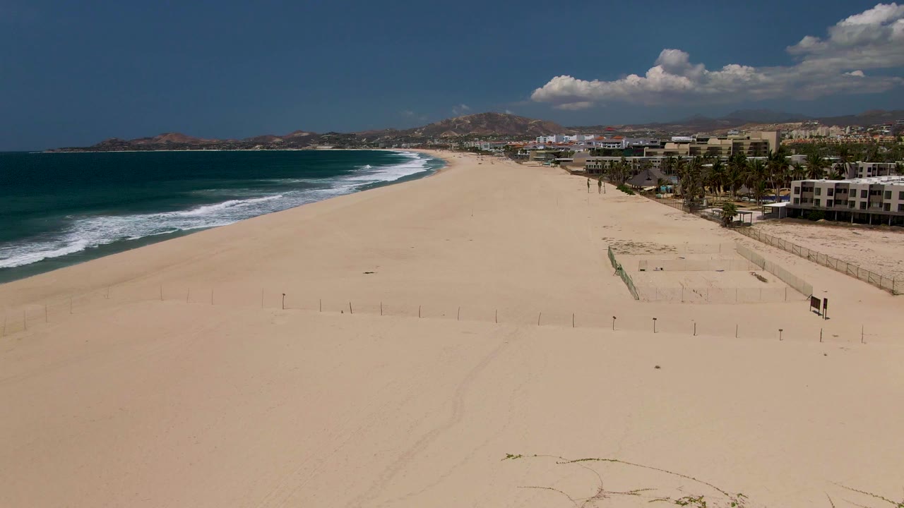 High view of the San Jose del Cabo coast with white surf bands meeting the sand, a fence line and buildings near the right edge, mountains in the distance, and no visible people.
