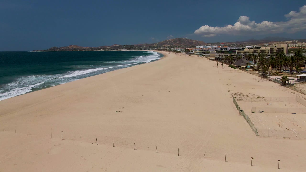 Opening aerial view of a wide San Jose del Cabo beach with blue ocean waves on the left, open sand in the foreground, a volleyball net to the right, distant buildings, palm trees, mountains, clouds, and tiny people visible.