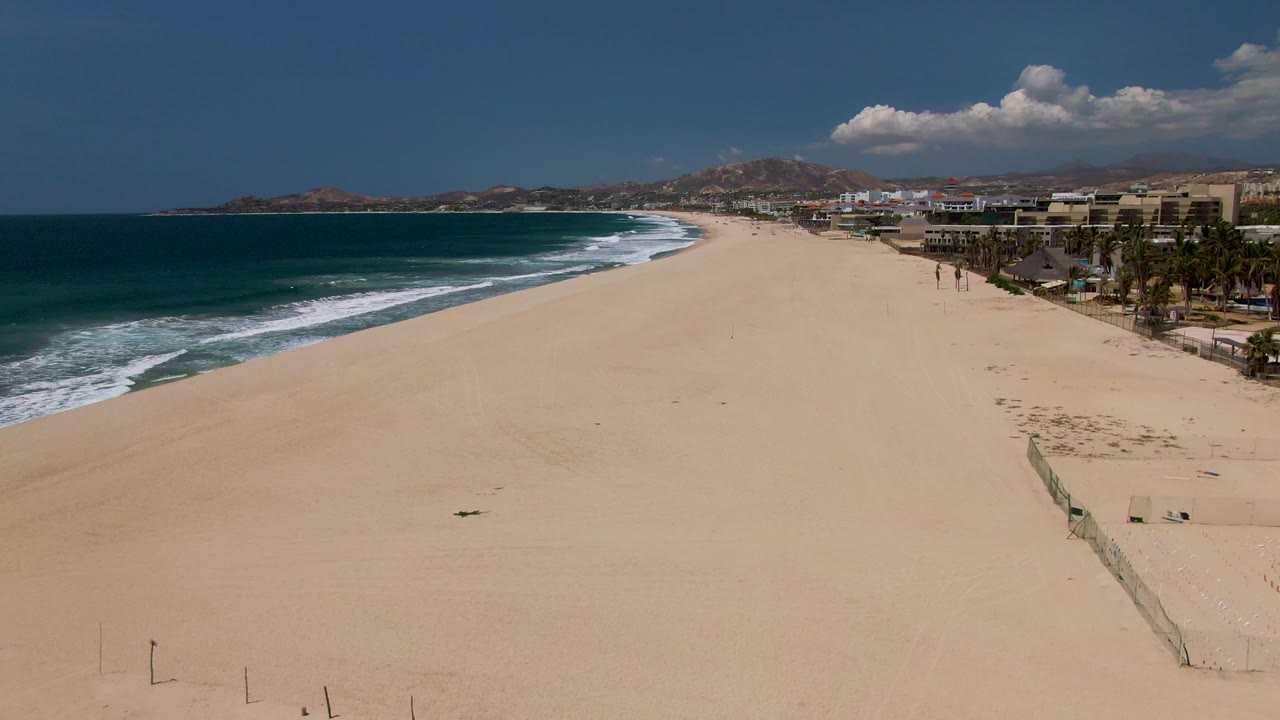 Sandy shoreline with blue water, surf, buildings, palms, and volleyball net on the right.