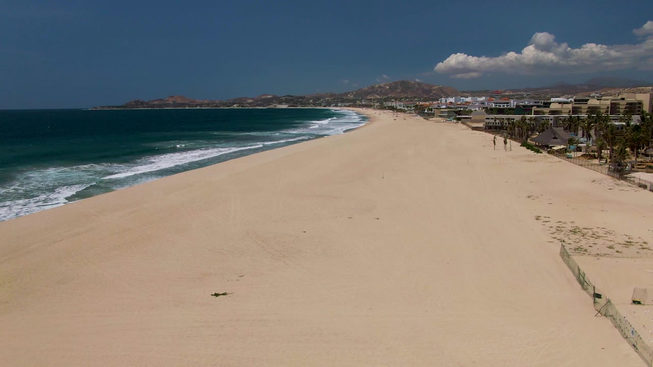 At 0:02, the aerial view shows the volleyball net sliding toward the right side of the San Jose del Cabo beach frame, with broad empty sand, white waves, buildings, palms, mountains, and small distant people.