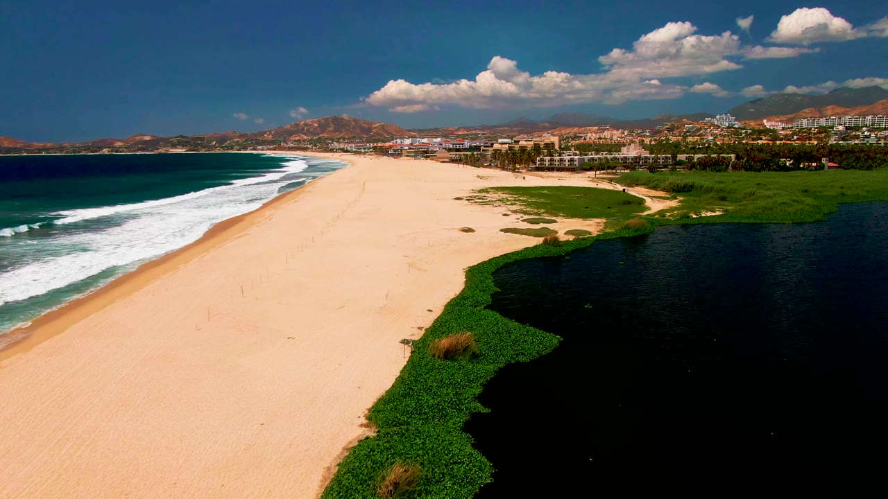 Aerial view of sandy beach, ocean surf, green vegetation, and dark lagoon water.