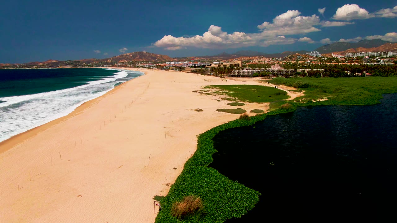 San Jose del Cabo coastline appears from above with white surf at left, broad sand through the center, green vegetation and black lagoon water at right, and no visible people.