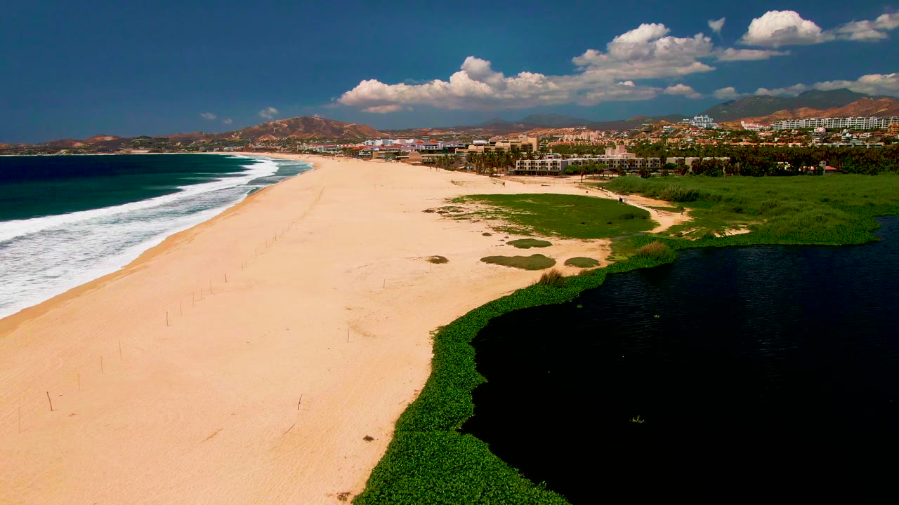 Aerial San Jose del Cabo beach view shows the surf line stretching along the left, empty sand in the middle, green dune vegetation and dark water on the right, and no visible people.
