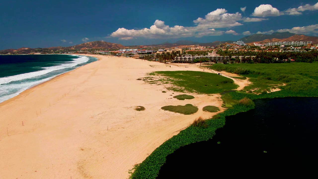 A wide aerial frame over San Jose del Cabo beach shows white surf on the left, open tan sand across the center, dark lagoon water and green vegetation on the right, distant buildings, hills, scattered clouds, and no visible people.