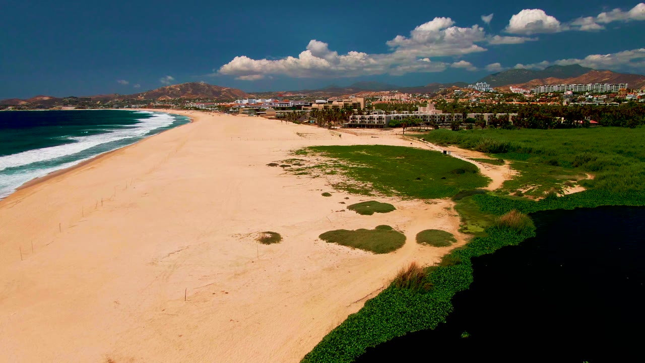 Aerial beach view with ocean waves, open sand, green lagoon edge, distant buildings, and hills.