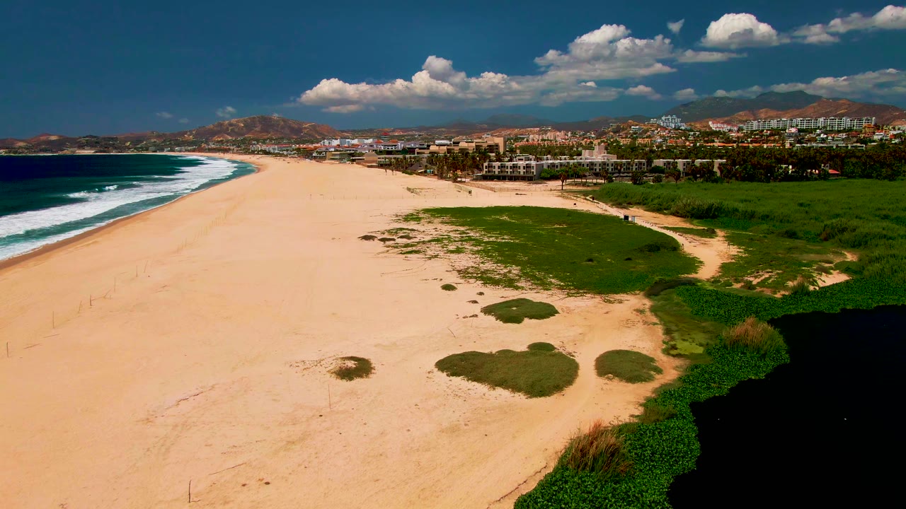 At 0:02, the aerial view emphasizes the open sand and green lagoon-side vegetation in the lower right, with white ocean surf to the left, San Jose del Cabo buildings in the distance, hills, clouds, and no visible people.