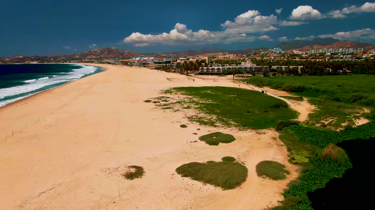 Aerial shoreline with broad sand, ocean waves, green patches, distant buildings, and hills.
