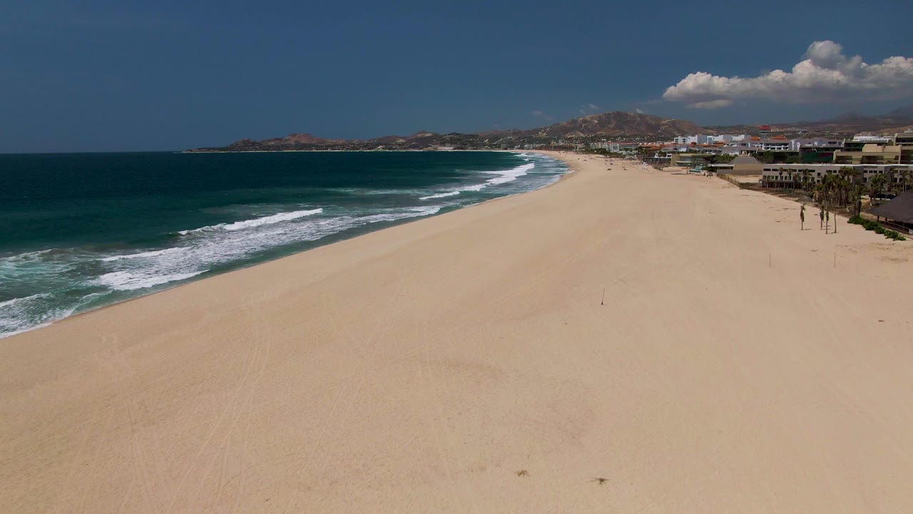 At 0:00, a wide aerial view in San Jose Del Cabo shows tan sand in front, dark blue ocean and white surf on the left, beachfront buildings on the right, and small people visible on the beach.