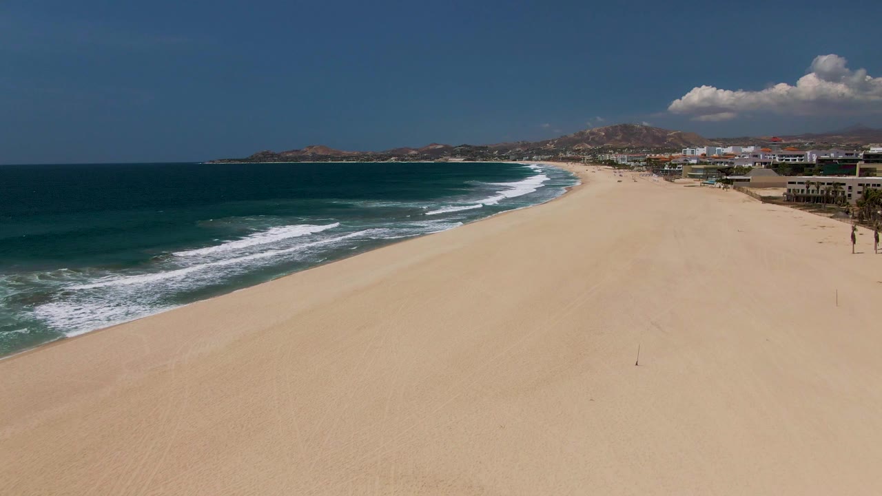 At 0:02, a high view over San Jose Del Cabo beach shows the surf edging closer across the left side, broad sand across the center, and low buildings and palms along the right background.