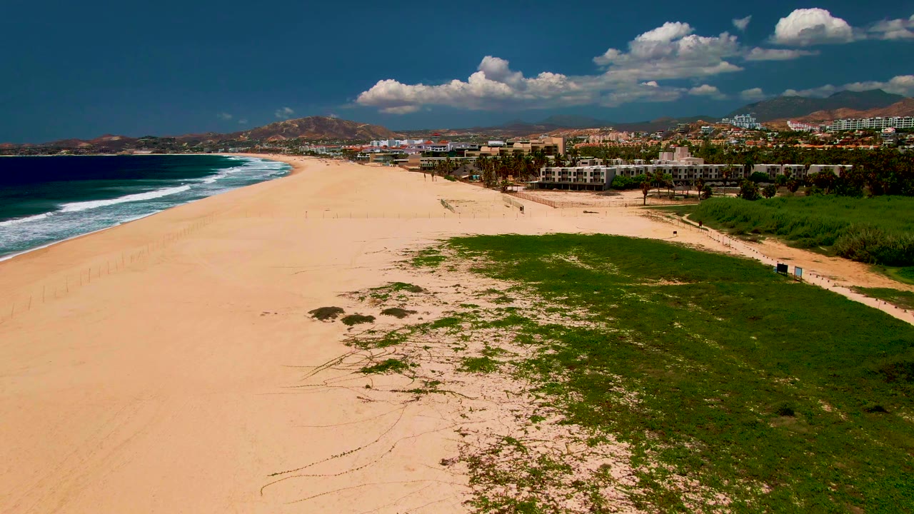 Aerial view over a San Jose del Cabo beach with white surf on the left, green dune vegetation in front, low buildings to the right, mountains beyond, and tiny people on the sand.