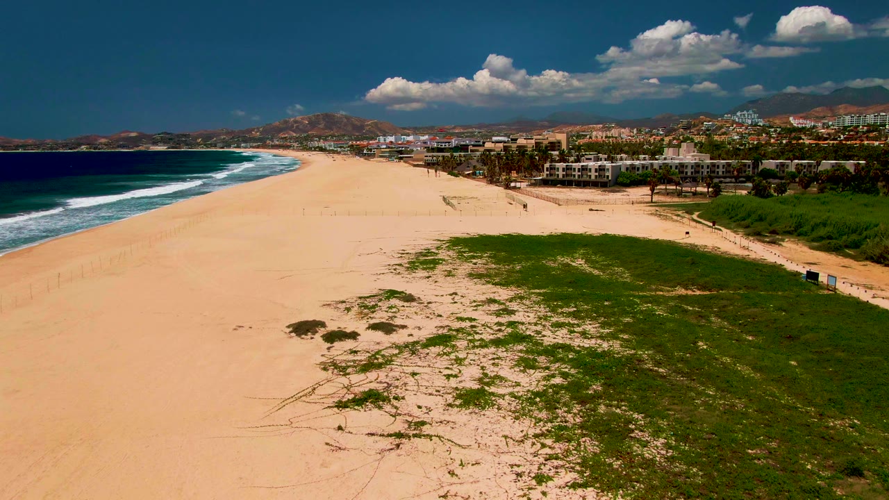 Wide drone view along beach with surf, dune grass, buildings, hills, and scattered clouds.