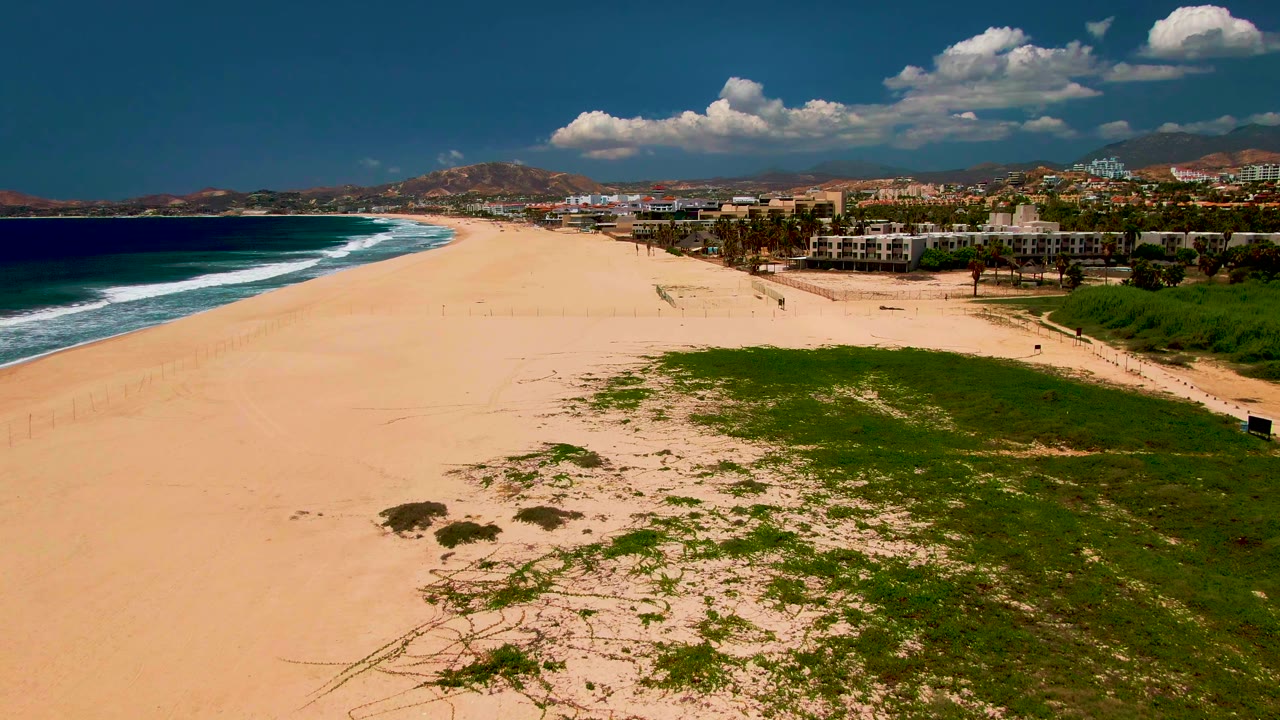 Aerial view in San Jose del Cabo with more open sand visible in the foreground, a patch of green dunes on the lower right, blue ocean and breaking waves at left, and distant people near the beach buildings.