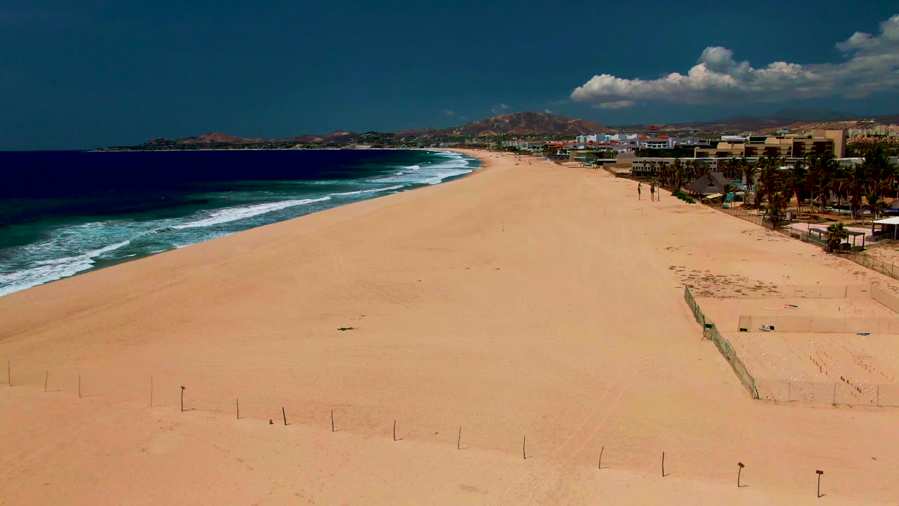 Wide opening frame over San Jose del Cabo beach with ocean waves on the left, broad sand in front, coastal buildings and palms on the right, distant mountains, and no visible people.