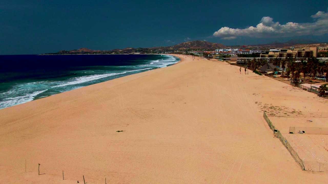One second into the clip, the San Jose del Cabo shoreline remains wide in frame, with turquoise surf advancing along the left, open sand across the center, buildings and palms to the right, and no visible people.