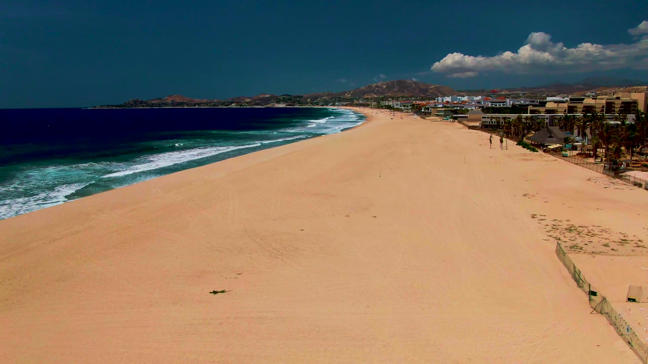 At 0:02, the San Jose del Cabo beach appears from a high diagonal angle, with blue ocean and white wave bands on the left, a large sand field, right-side buildings and palms, and no visible people.