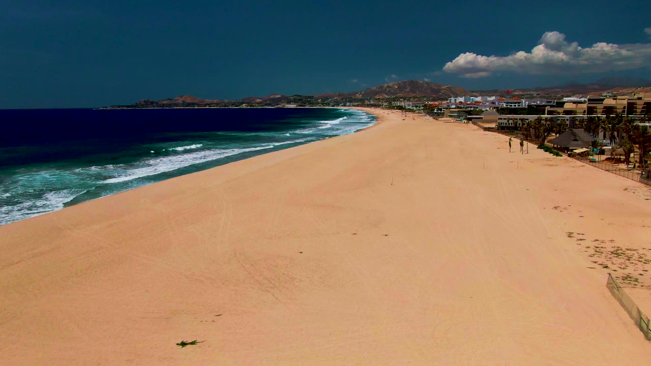 At 0:03, the aerial view looks down the San Jose del Cabo coastline, with darker blue water at left, breaking surf, empty sand, beachside structures and palms at right, and no visible people.