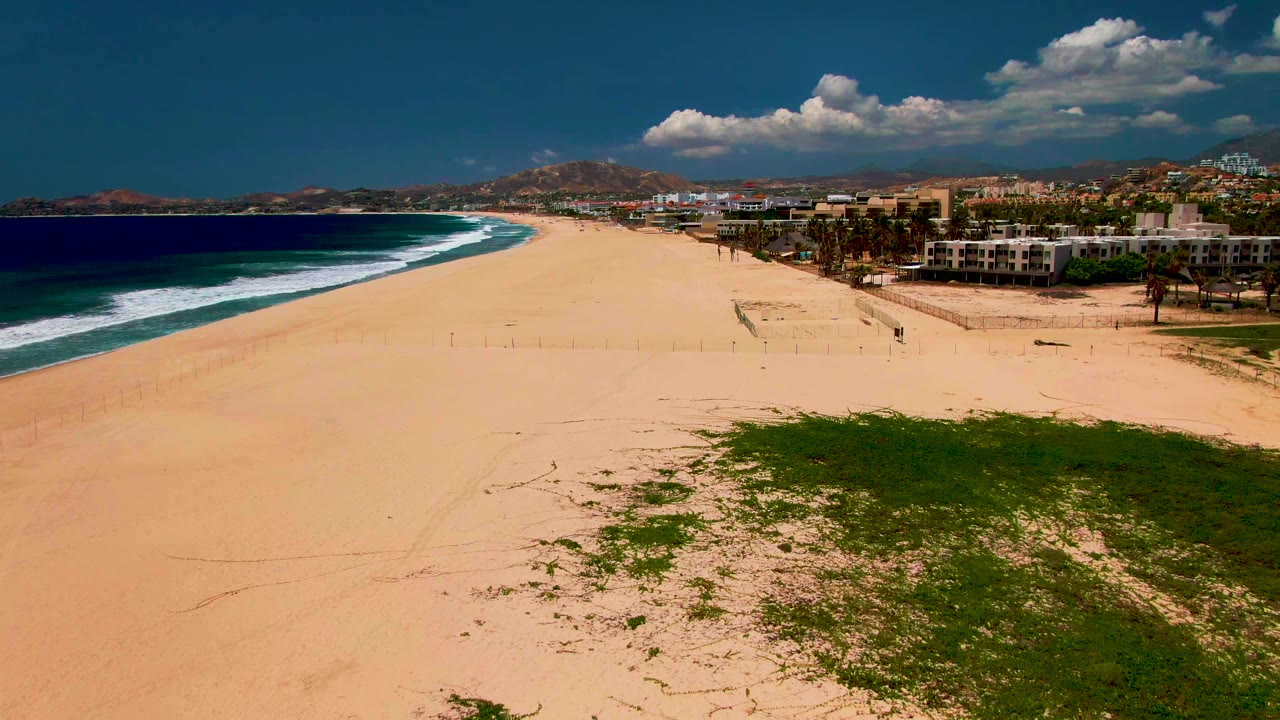 Opening elevated view in San Jose Del Cabo showing tan beach, blue ocean surf on the left, green dune vegetation in the foreground, coastal buildings to the right, and no visible people.
