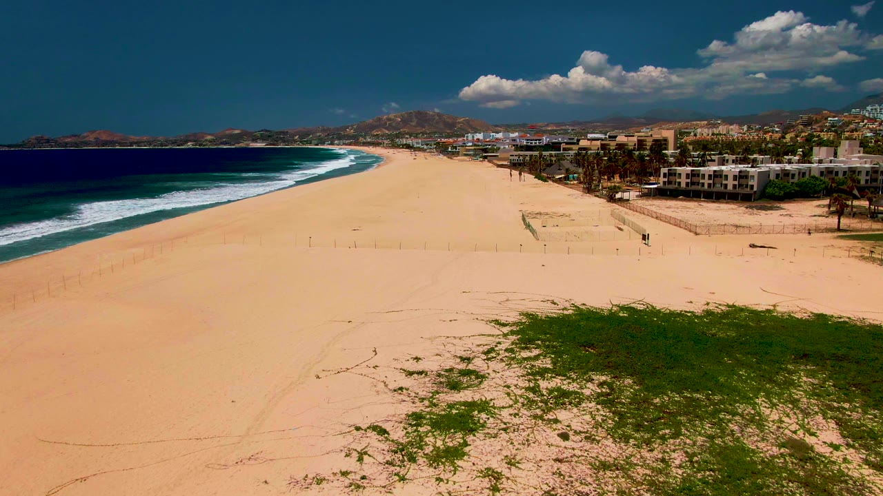 San Jose Del Cabo beach frame with the surf line curving along the left edge, broad open sand in the center, beachfront buildings on the right, and no visible people.