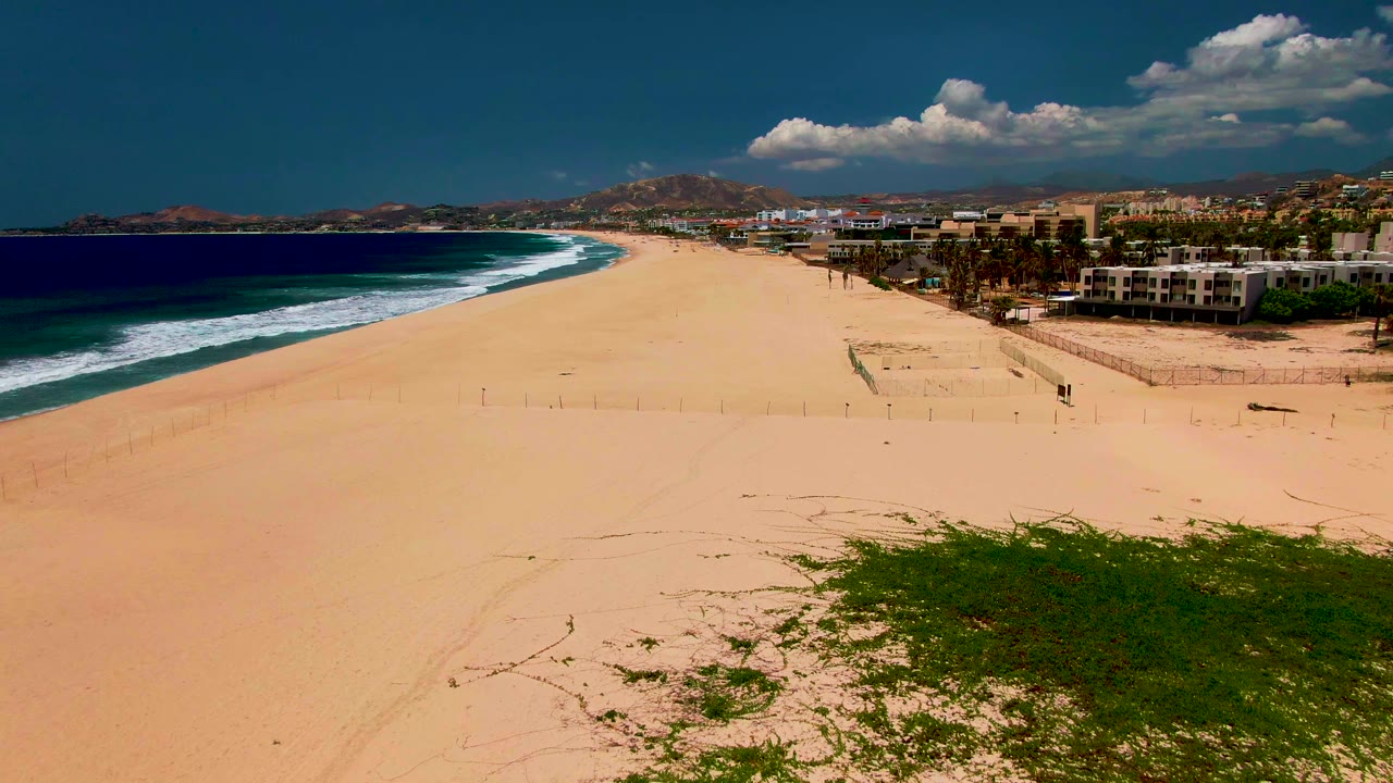 Elevated San Jose Del Cabo shoreline view with more open sand in the foreground, dark blue water to the left, low buildings and palms to the right, and no visible people.