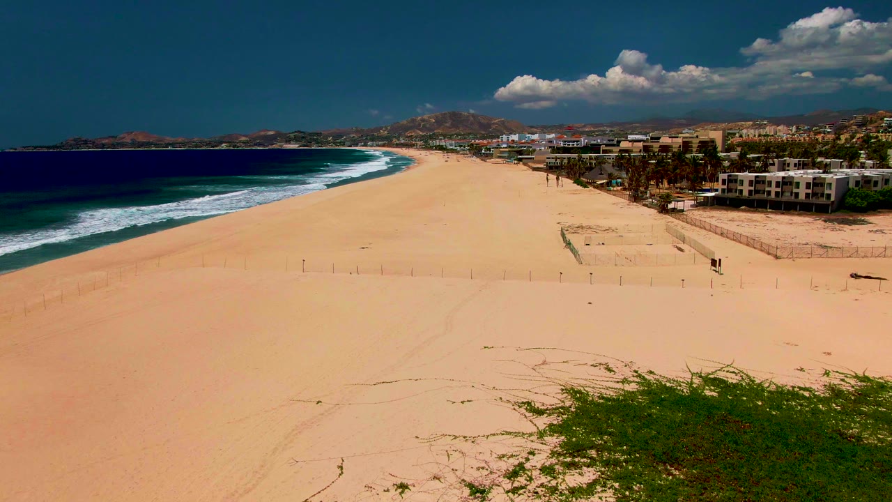 Aerial beach view with surf, pale sand, coastal buildings, and hills in the distance.