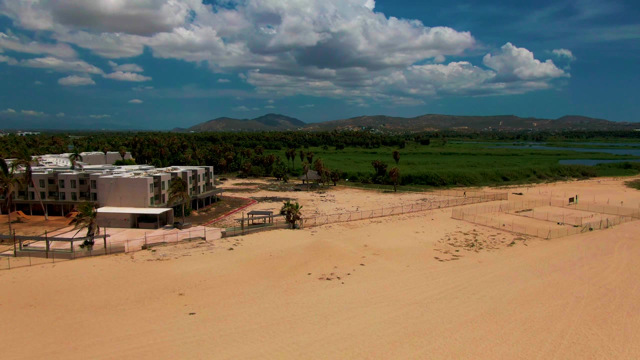 Aerial frame over San Jose del Cabo keeps the building block on the left side while broad sand fills the foreground and green fields, water, hills, and cloud cover stretch across the background with no visible people.