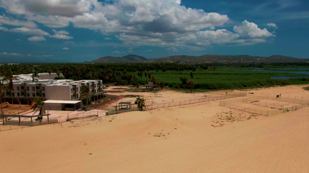 The San Jose del Cabo aerial view shows the buildings slightly farther left, a fenced sandy area across the midground, palms near the structures, green land and water behind, and no visible people.
