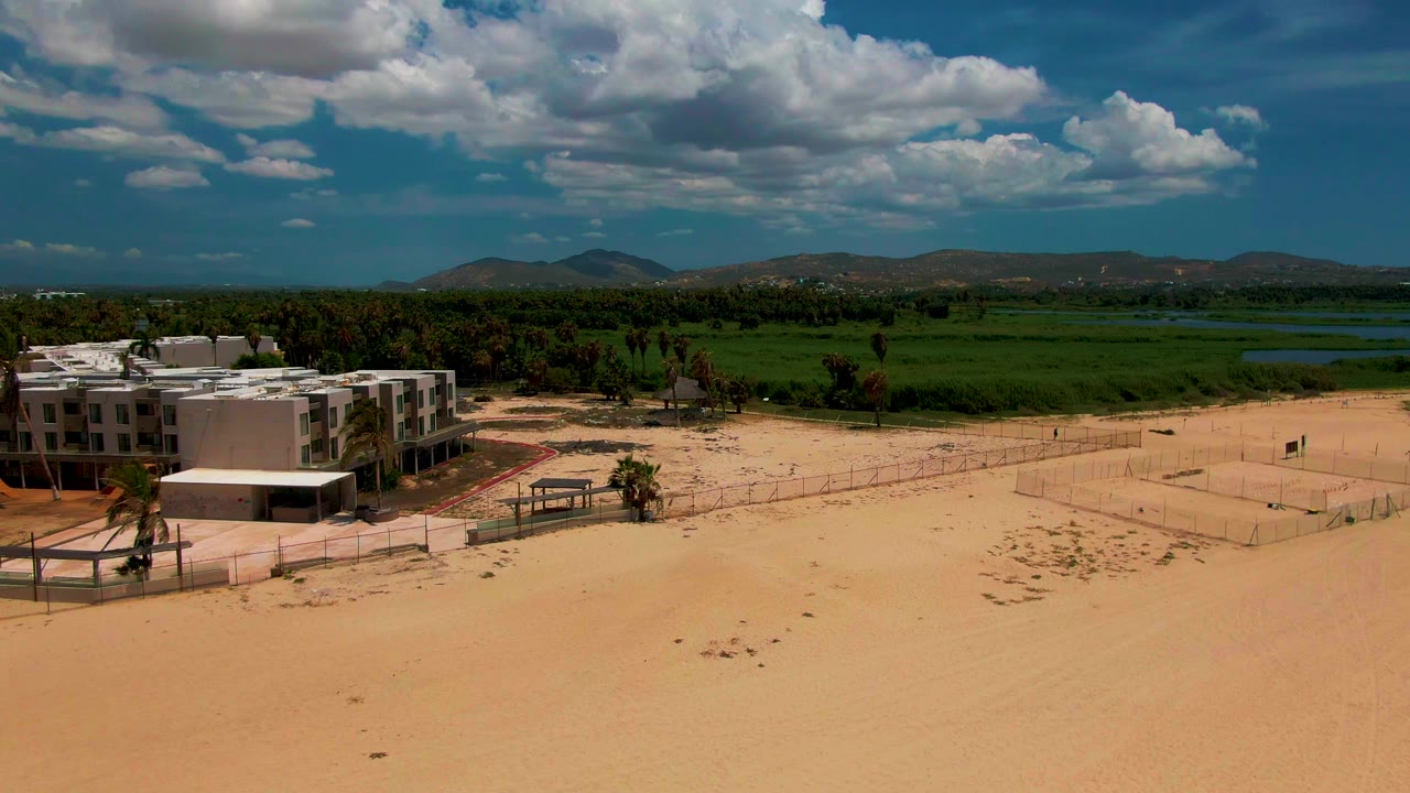 Mid-clip aerial frame in San Jose del Cabo places the buildings partly cropped at the left edge, with open sand below and a broad green field, water, hills, blue sky, and no visible people behind it.