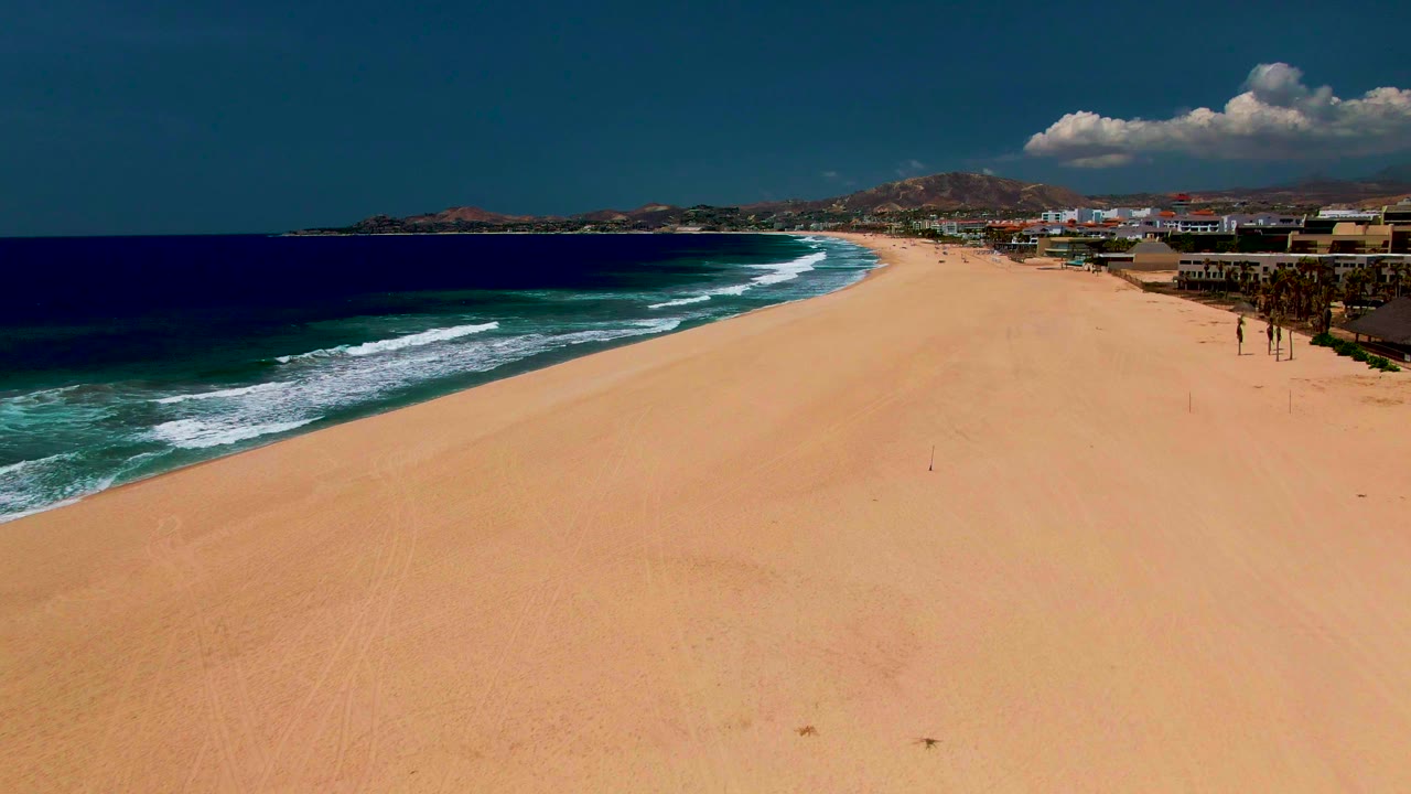Wide aerial view of a sandy beach in San Jose del Cabo with dark blue ocean on the left, white surf lines, oceanfront buildings on the right, distant hills, clouds, and small people on the sand.