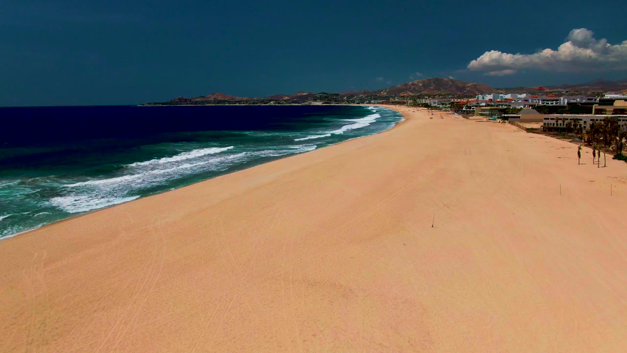 Sandy beach and curving surf beside oceanfront structures and hills.