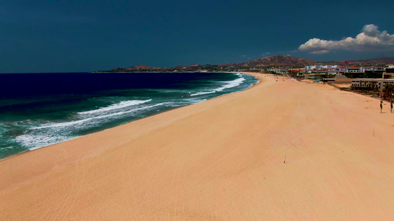 The San Jose del Cabo beach remains wide and open as the surf forms diagonal white lines at left, tan sand fills the center, buildings sit along the right edge, and tiny people are visible near the shoreline area.