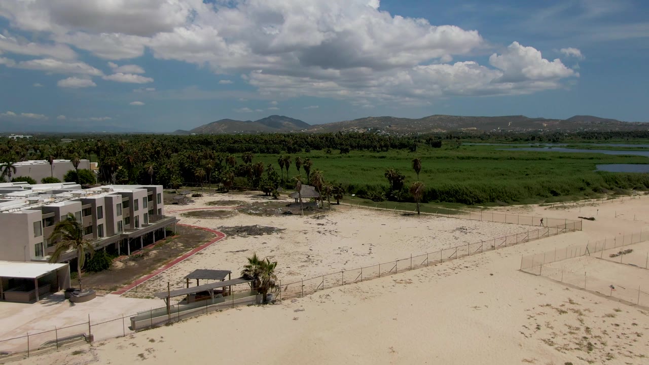 At 0:00 in San Jose del Cabo, a white resort building sits at the left edge of a sandy beach with palms, fencing, green fields, distant hills, blue sky, white clouds, and no visible people.